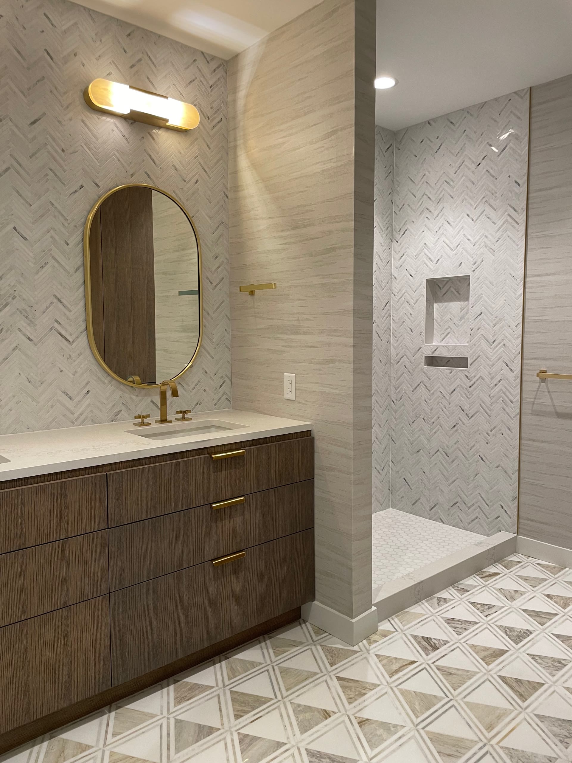 Floating wood vanity with oval mirror beside a herringbone-tiled shower