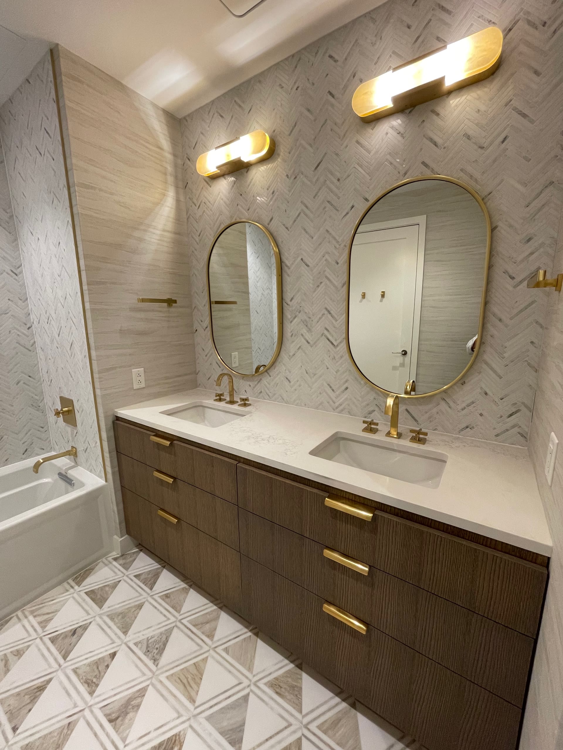 Double vanity with herringbone marble backsplash, oval mirrors, and brass hardware